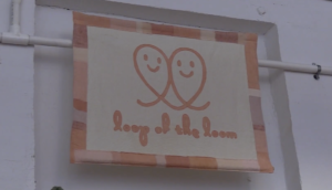 Two loops with smily faces in their centers come close to one another, as if hugging, with the words "loop of the loom" written beneath them in cursive, to form the company Loop of the Loom's logo